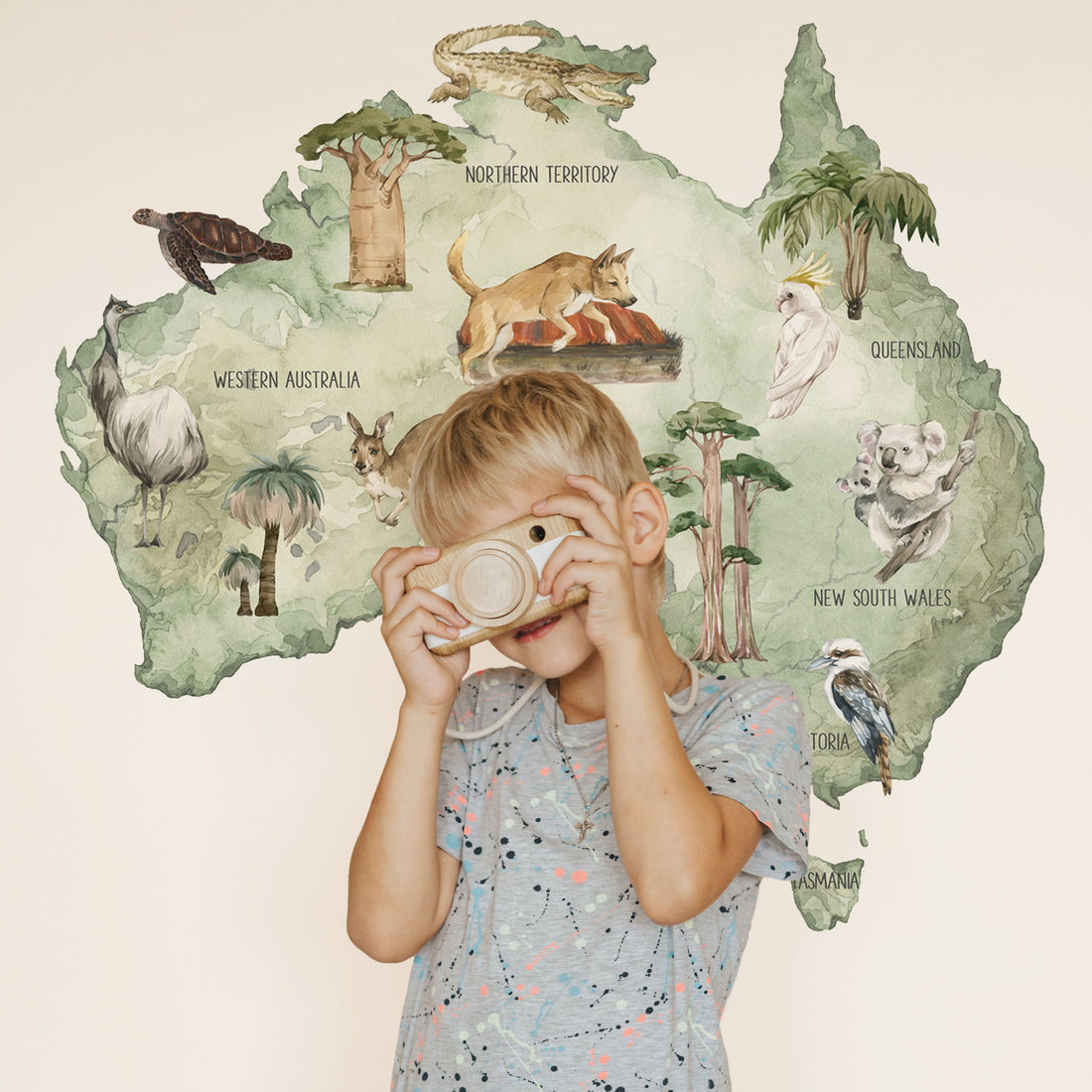 A young child stands in front of a large wall mural decal shaped like Australia, The wall decal features illustrated native animals and landmarks on a pale sage green background.