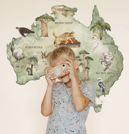 A young child stands in front of a large wall mural decal shaped like Australia, The wall decal features illustrated native animals and landmarks on a pale sage green background.