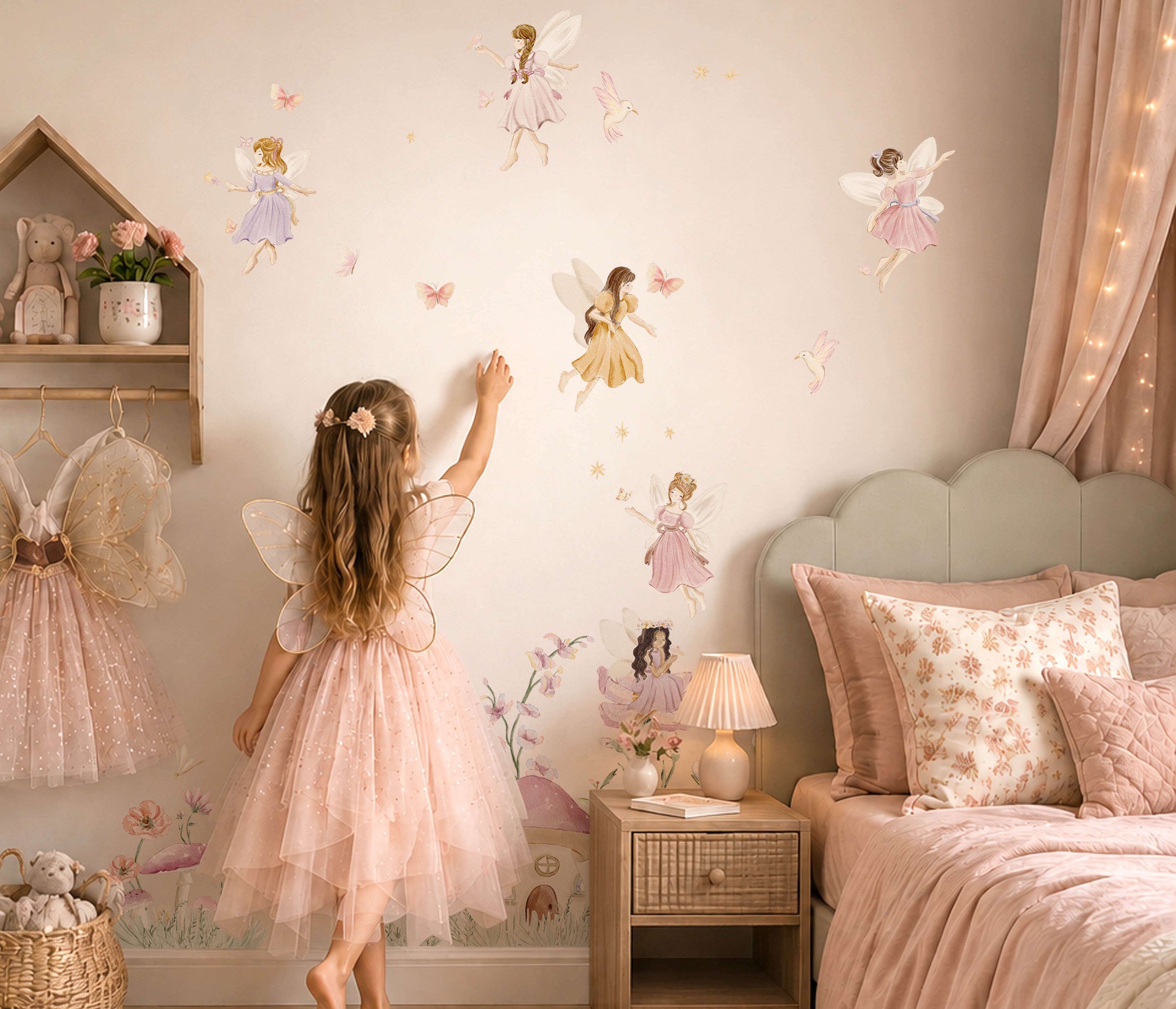 A young girl in a pink fairy costume stands in a cozy, fairy-themed bedroom, touching a wall decorated with large fairy decals. The room features a matching pink bed, a wooden nightstand, a small house shelf with fairy dresses, and warm fairy lights.