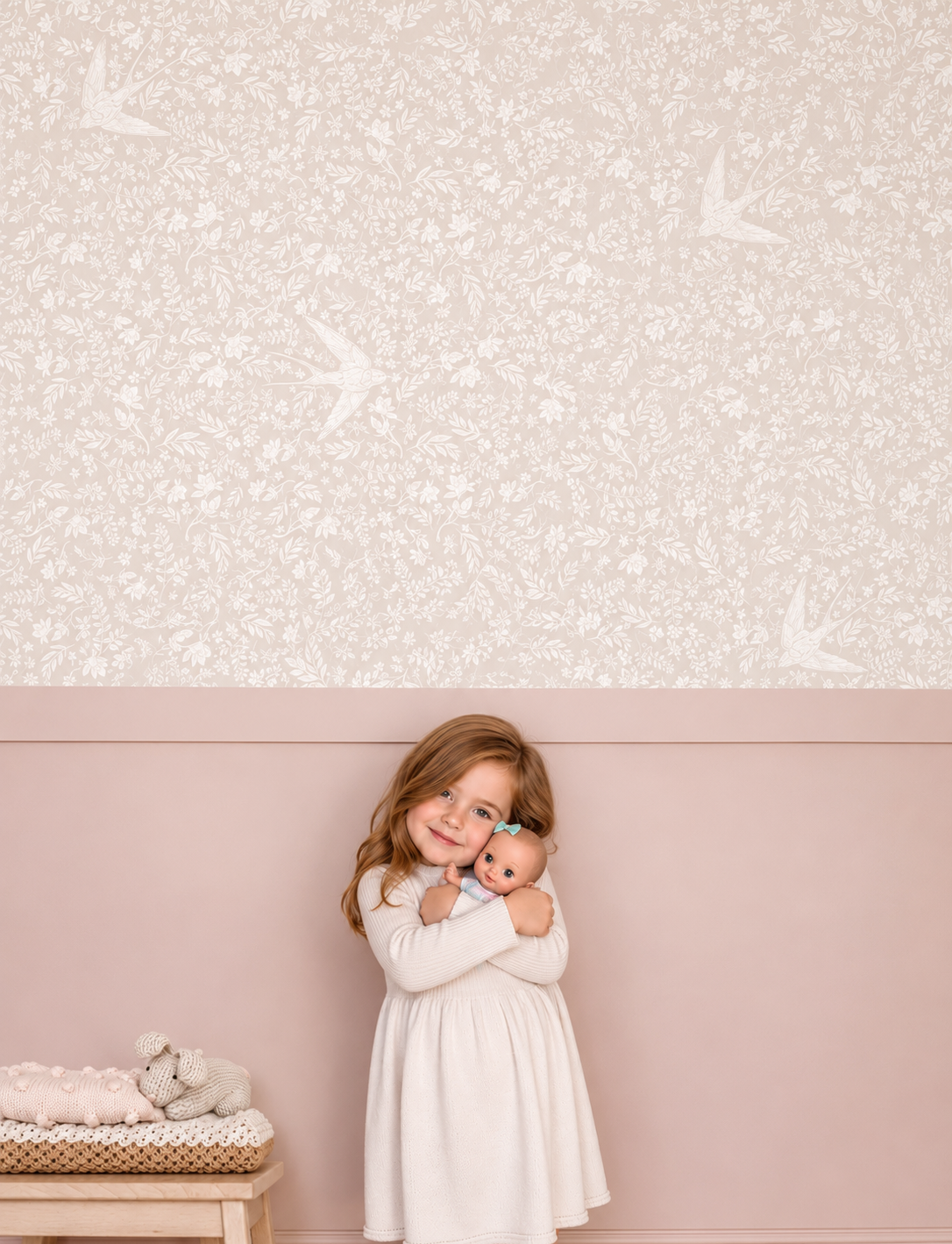 A young girl with long light brown hair stands against a two-tone wall, holding a doll. The wall above her has a light pink floral and bird pattern, and the lower wall is dusty pink.