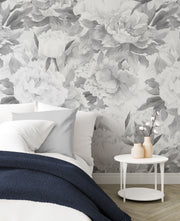 A styled bedroom corner featuring a large-scale grayscale floral wallpaper with peony-like flowers. A bed with white and gray pillows and a navy blue throw blanket is partially visible, alongside a small white side table with two beige vases.